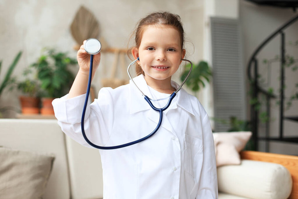 Hymyilevä pieni tyttö lääketieteellisessä univormussa leikkii stetoskoopilla kotona - Valokuva, kuva