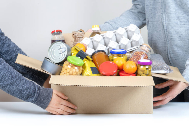 Volontari mani in possesso di scatola di donazioni di cibo con prodotti alimentari sulla scrivania bianca - Foto, immagini