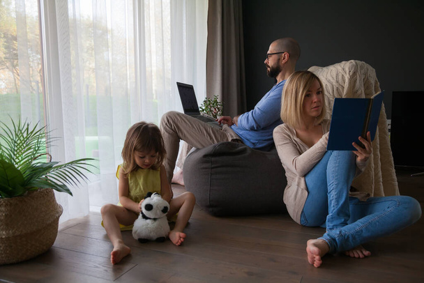 Família passando tempo juntos em casa pai com laptop, mãe lendo um livro e sua filhinha brincando sozinha com seu brinquedo - Foto, Imagem
