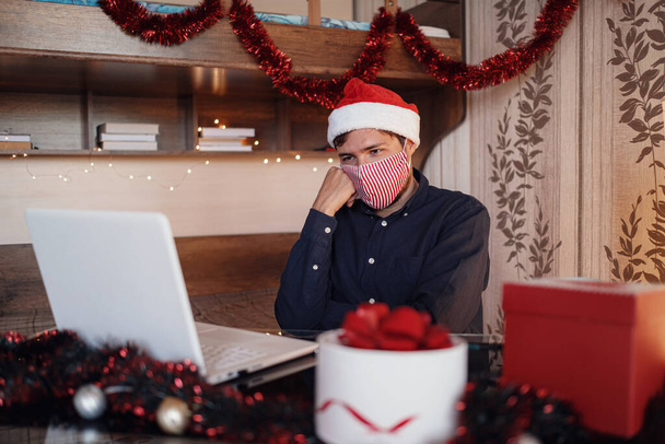 クリスマスの装飾の近くの部屋に座っている仮面の不幸な男の肖像画光と贈り物を注文する。新年のeショッピング。メリークリスマスCovid 19コロナウイルスの社会的距離の概念. - 写真・画像