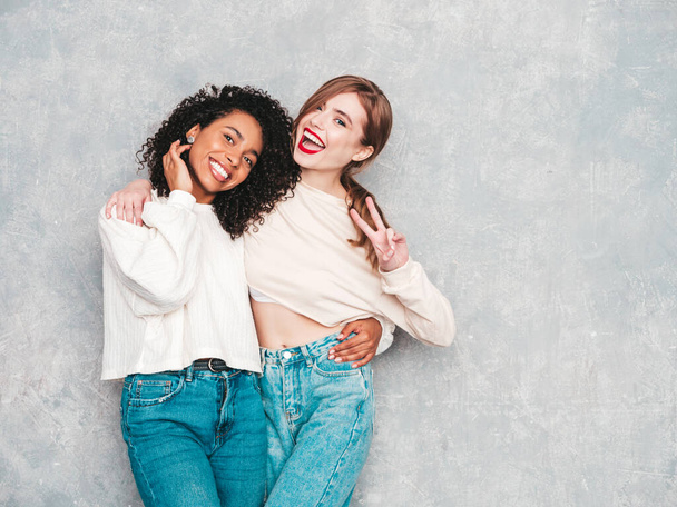 Two young beautiful smiling international hipster female in trendy summer jeans clothes. Sexy carefree women posing near gray wall in studio. Positive models having fun. Concept of friendship  - Valokuva, kuva