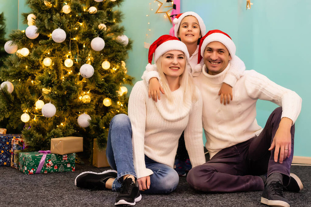 No Natal. Família. Felicidade. pai, mãe e filha em chapéus de Papai Noel olhando para a câmera e sorrindo no chão em casa - Foto, Imagem