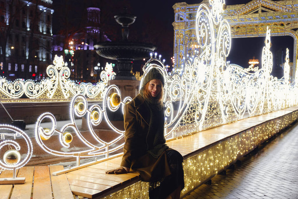 路上でポーズしている若い笑顔の女性。背景にあるお祭りのクリスマスフェア。スタイリッシュな冬のコート、ニットキャップ、スカーフを身に着けているモデル。大都市で幸せな気分。冬の休暇を過ごすモスクワ - 写真・画像
