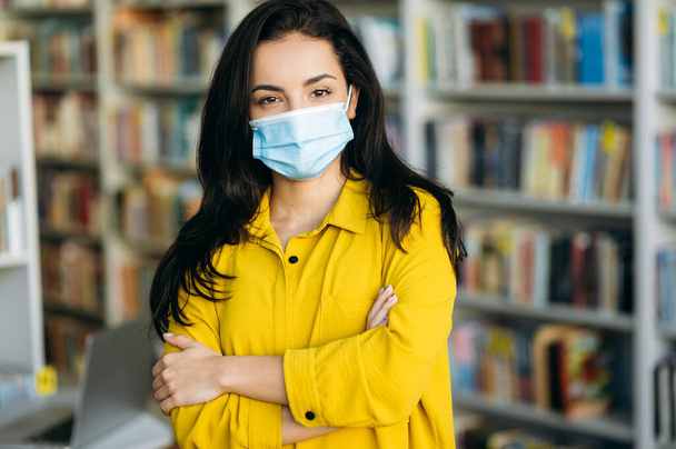 Close-up portrait of caucasian beautiful woman in casual yellow shirt standing indoors with arms crossed in front wearing a protective medical mask on her face and looking directly into the camera - Foto, Imagen