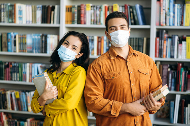 Tarz sahibi genç bir erkek ve kadın, koruyucu maskeler takan öğrenciler kütüphanede el ele kitap tutarak kameraya, sağlık hizmetleri konseptine bakıyorlar. - Fotoğraf, Görsel