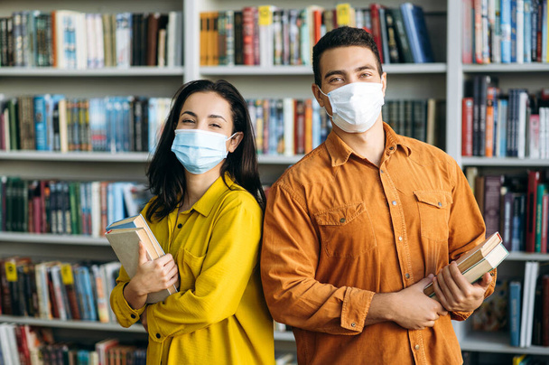 Modern öğrenciler, koruyucu yüz maskeleri takıp kütüphanede yanyana duruyorlar ve ellerinde kitaplarla kameraya bakıyorlar, sağlık sigortası konseptine. - Fotoğraf, Görsel
