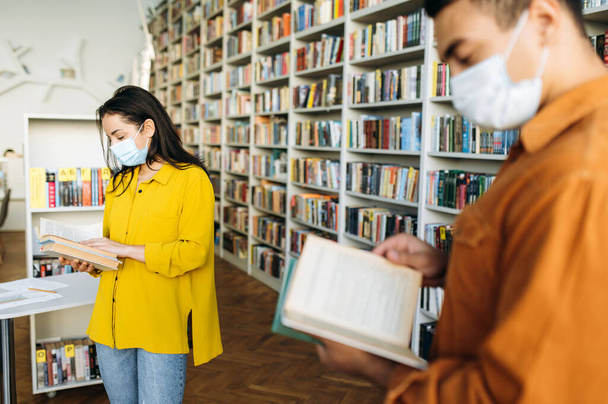 Learning during pandemic. Stylish students, girl and guy, wearing protective face masks stand at a distance from each other in the library and reading books, healthcare concept - Fotografie, Obrázek