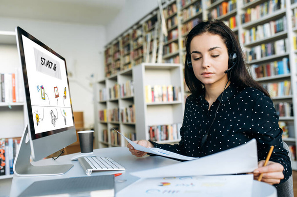 Young successful woman with headset sitting at her desk and develops a startup strategy by studying financial graphs - 写真・画像