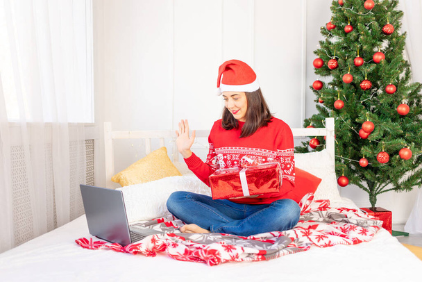 a young beautiful girl in a red sweater and a hat is making a video call with a gift on a laptop at home on a bed near a Christmas tree with a new year's interior - Fotó, kép