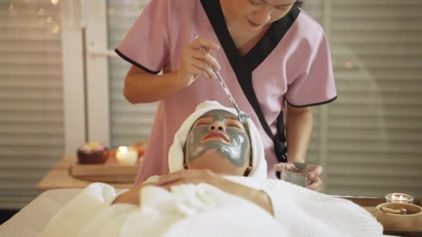La masajista en el spa utiliza un cepillo para aplicar una máscara de barro a una hermosa mujer asiática - Imágenes, Vídeo