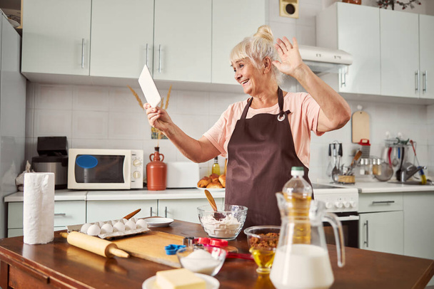 Fröhliche ältere Dame macht Videoanruf aus ihrer Küche - Foto, Bild