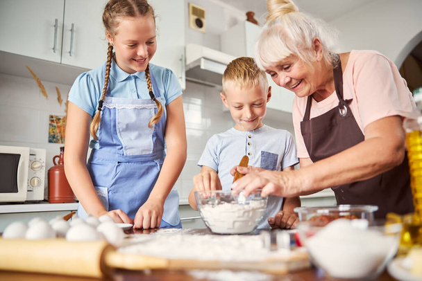 Friendly senior lady showing her grandchildren how to make dough - Zdjęcie, obraz