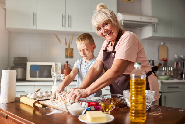 Χαρούμενη ηλικιωμένη κυρία και εστιασμένο αγόρι μαγείρεμα στην κουζίνα - Φωτογραφία, εικόνα