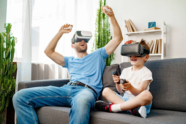 Dad and son sitting on sofa and playing games with VR glasses - Foto, Imagem