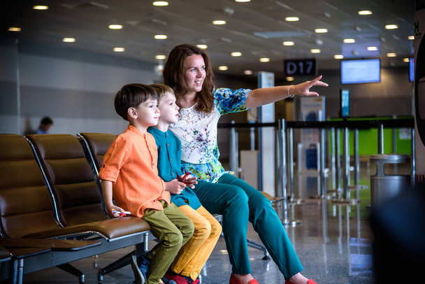 Matka i dwaj bracia syn wskazujący na startowanie samolotów przez szybę okienną na lotnisku czekając na ich lot. - Zdjęcie, obraz