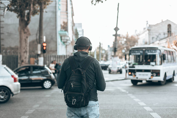 Visão traseira do homem em roupas casuais com uma mochila ouvindo música em fones de ouvido na rua ao ar livre. - Foto, Imagem