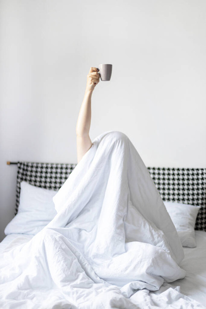 Woman showing arm raised up holding coffee cup on bed in bedroom - Фото, зображення