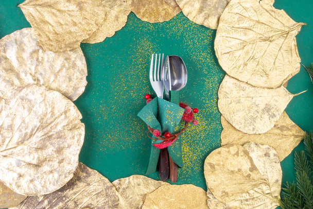 Kerst bestek ingesteld op trendy groene achtergrond met goudkleurige bladeren. Creatieve feestelijke tafelschikking. Vlakke lay, bovenaanzicht, kopieerruimte. - Foto, afbeelding