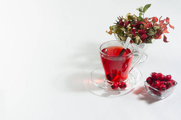 Organic red hawthorn tea in transparent cup, fresh hawthorn berries, rose hip in sunlight with shadow on white wood board, copy space. - Zdjęcie, obraz