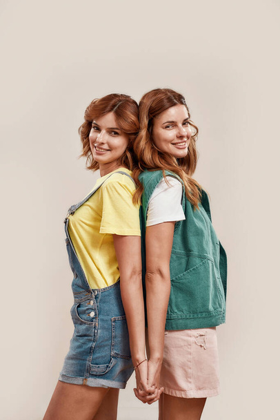Portrait of two attractive young girls, twin sisters in casual wear smiling at camera, holding hands, posing together, standing back to back isolated over light background - Foto, Bild