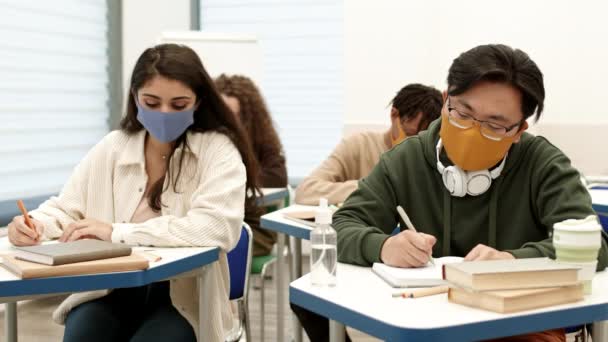 Mixed-Race College Studentin sitzt am Schreibtisch im Klassenzimmer neben asiatischen männlichen Klassenkameraden, kopiert seine Vorlesung, er bemerkt und bedeckt Notizbuch, Mädchen kichern, Menschen mit Mundschutz - Filmmaterial, Video