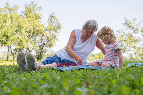 Babička s vnučkou ležérně sedí na dece a čtou a povídají si v parku. - Fotografie, Obrázek
