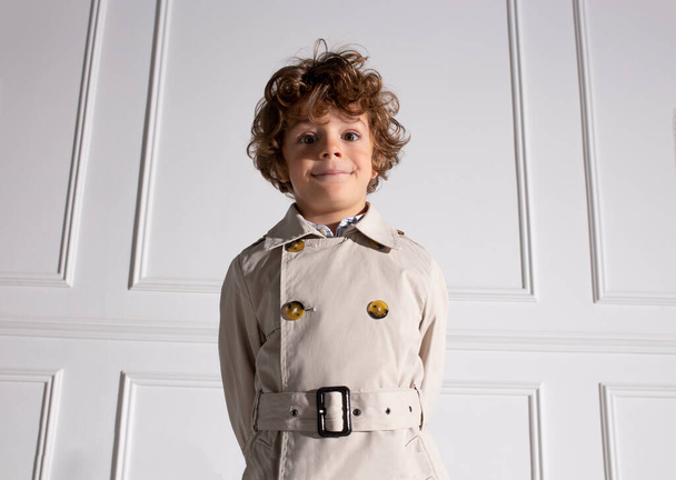 Closeup portrait of a curly little boy, wear in trench, poses in studio, over white background. - Foto, Imagem