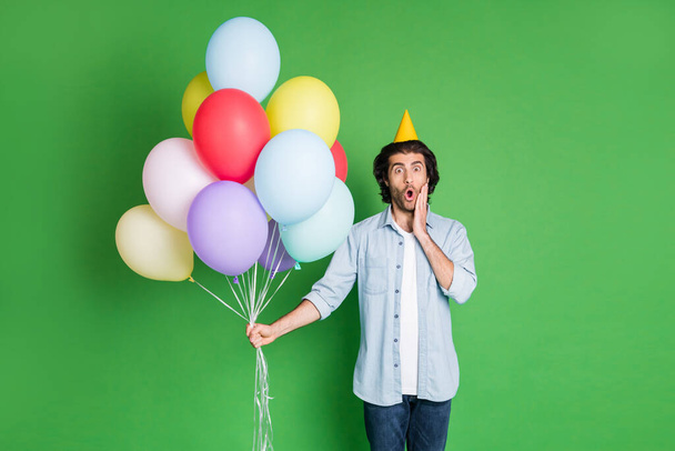 Photo of young bearded guy hold balloons open mouth hand cheek wear paper cone hat denim shirt jeans isolated green color background - Foto, immagini