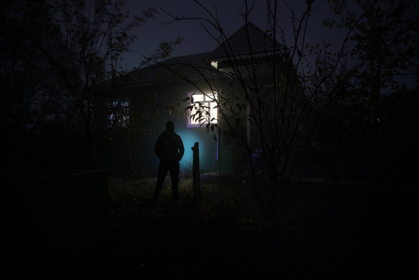 Stary dom z Duchem w lesie w nocy. Przerażająca sylwetka przy oknie. Horror Halloween koncepcja Samotny człowiek sylwetka stoi w pobliżu okna domu w nocy. Skupienie selektywne - Zdjęcie, obraz