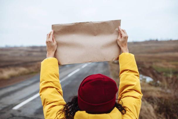 Backview de uma turista mulher vestindo casaco amarelo e chapéu vermelho fica com um papel em branco cartaz no lado de uma estrada de inverno vazio. Copyspace, lugar para o seu texto. Hitch-hiking, conceito de viagem - Foto, Imagem