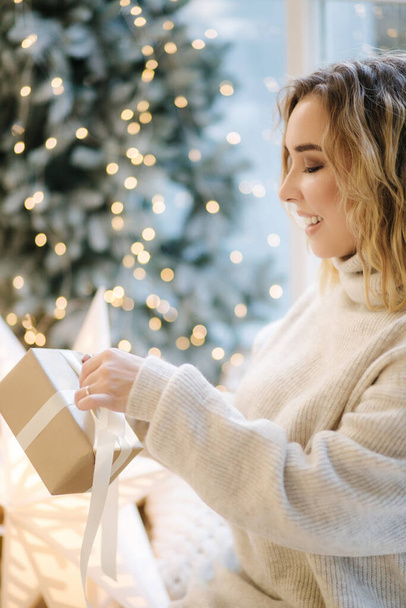 Portrait of blond hair woman in hoodie open present on Chrisrmas time. Elegant woman stand by big Christmas tree in front of window - Photo, Image