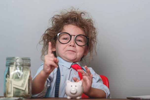 Funny little kid investor with finger poiting up and biggy bak, calculator and jar with money. Child in suit looks like a businessman. - Foto, imagen