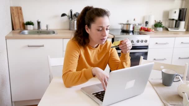 Femme faisant du shopping en ligne et payant par carte de crédit en or. Jeune fille assise avec ordinateur portable achetant sur Internet entrez les détails de carte de crédit sur fond de cuisine intérieure. Boutique en ligne e-commerce concept - Séquence, vidéo