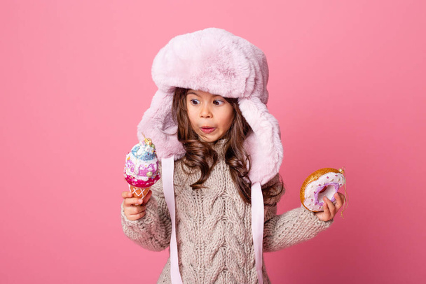 κοριτσάκι με ζεστό καπέλο με χριστουγεννιάτικα στολίδια σε σχήμα παγωτού και ντόνατ - Φωτογραφία, εικόνα