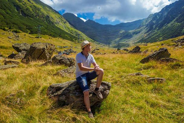 Man in straw hat sits on a rock against the background of high mountains. Hiker sitting on the cliff edge near Transfagarasan, one of the most spectacular mountain road, best according to Top Gear - Photo, Image