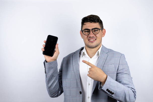 Jeune homme d'affaires portant une chemise décontractée sur fond blanc montrant et pointant son téléphone - Photo, image