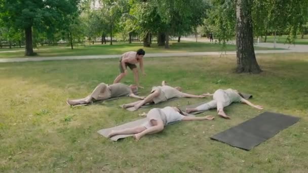 Amplia toma de cuatro mujeres ancianas y un hombre acostado en colchonetas de yoga en la hierba y estirando sus cuerpos con la ayuda de un entrenador femenino de mediana edad - Metraje, vídeo