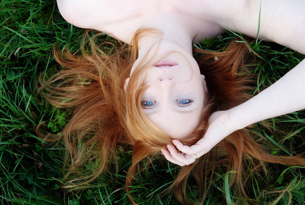 Portrait of a beautiful young sexy redhead woman spreading red hair fan-shaped lying with bare shoulders relaxing on the green grass, upside down, copy space - Foto, Imagen