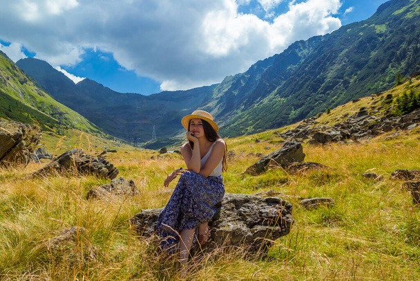 Dívka v šatech a slaměný klobouk sedí na skále na pozadí vysokých hor. Žena na okraji útesu v blízkosti Transfagarasan, jedna z nejkrásnějších horských silnic, nejlepší podle Top Gear - Fotografie, Obrázek