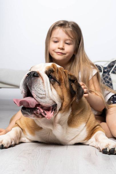 Egy fiatal lány a hálószobában, ül a földön a kutyájával és simogatja. Gyerek és kutya. Az angol bulldog egy fajtiszta kutya, származású.. - Fotó, kép