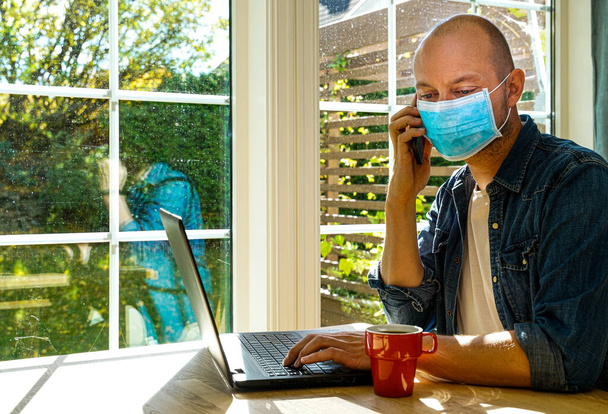 Homem adulto usando uma máscara facial e sentado na frente do laptop, fala em um telefone celular enquanto rola no PC. .. Foto de alta qualidade - Foto, Imagem