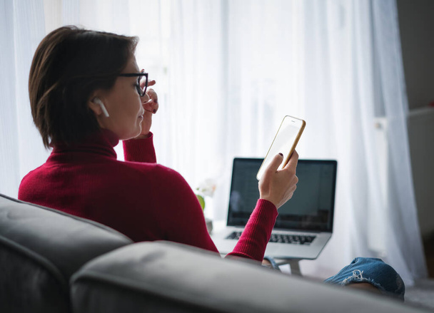 Dorosła kobieta pracuje w domu online trzymając telefon wideo czat z laptopem duże okno w tle w salonie - Zdjęcie, obraz