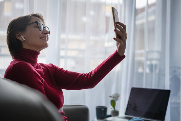 Mujer adulta que trabaja en casa en línea celebración de chat de vídeo de teléfono con el ordenador portátil gran ventana en el fondo en la sala de estar - Foto, imagen