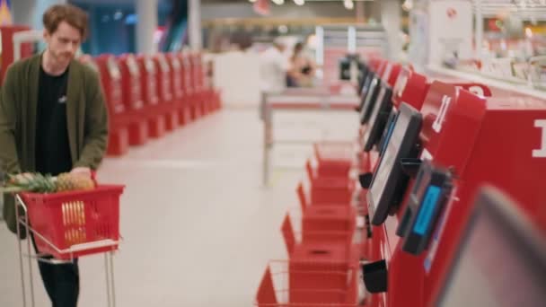Średni ujęcie młodego człowieka z koszyka zakupu produktów żywnościowych przy użyciu systemu self-checkout skanowania przedmiotów w hipermarkecie - Materiał filmowy, wideo
