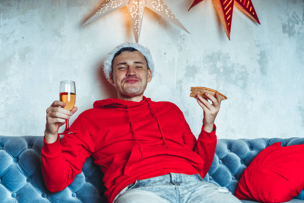 Young man in santa hat drinks champagne and eats pizza, sitting on sofa in room. Happy male resting with food and alcohol. Concept of Christmas celebration at home - Photo, Image