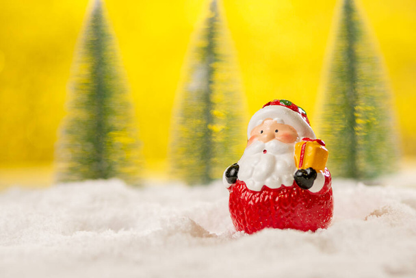 Keramiek figuur van de Kerstman in de sneeuw tussen kerstbomen op een abstracte gele achtergrond. - Foto, afbeelding