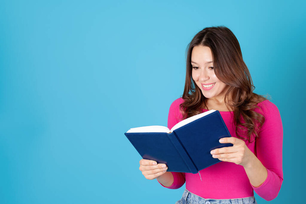 close-up portrait of a smiling young woman holding a diary or book in her hands and reading it with pleasure, isolated on a blue background - Photo, image