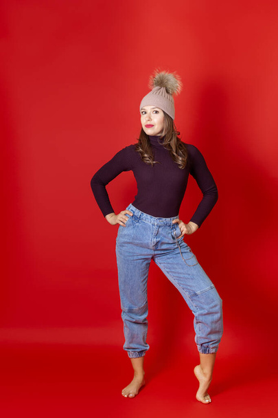 a full-length smiling young woman stands in a knitted winter hat and jeans, isolated on a red background - Fotografie, Obrázek