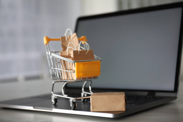 Internet shopping. Modern laptop and small cart with bags and box on table indoors - 写真・画像