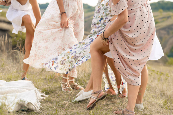 The company of female friends having fun, dancing on summerfield in long dresses showing their legs. Summer rural style picnic concept - Фото, изображение
